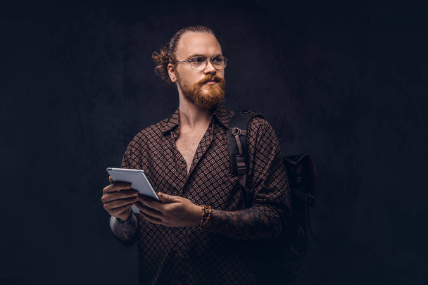 Portrét zrzka bokovky studenta v brýlích oblečený v hnědé tričko, má batoh a digitální tablet, pózuje ve studiu. Izolované na tmavém pozadí. - Fotografie, Obrázek