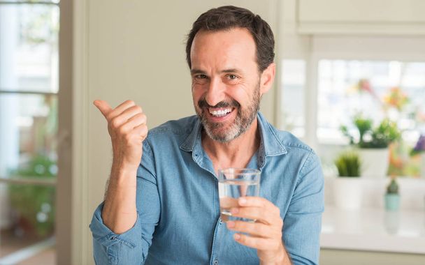 Чоловік середнього віку п'є склянку води, вказуючи рукою і пальцем вгору з щасливим обличчям посміхаючись
 - Фото, зображення