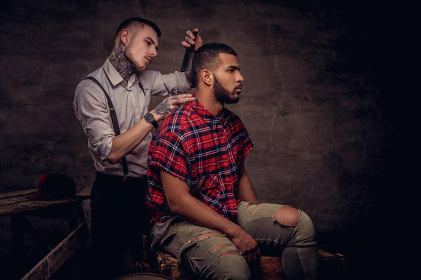 Homem afro-americano bonito recebendo um corte de cabelo enquanto sentado em caixas de madeira em um estúdio. Cabeleireiro tatuado profissional à moda antiga faz um corte de cabelo
. - Foto, Imagem
