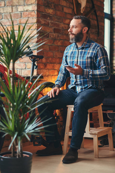 schöner bärtiger Hipster-Mann in blauem Fleece-Hemd und Jeans hält ein Smartphone in der Hand, während er auf einem Holzhocker in einem Studio mit Loft-Interieur sitzt. - Foto, Bild