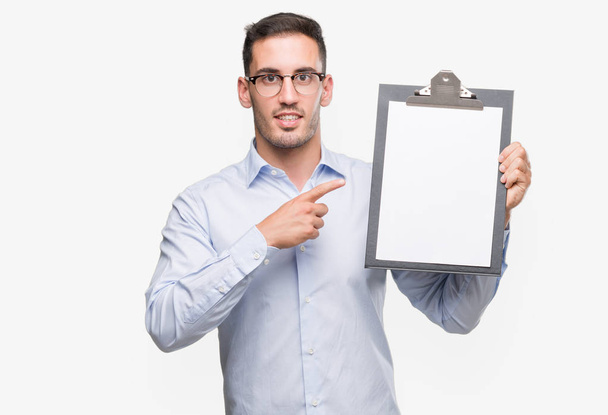 Handsome young business man showing a clipboard very happy pointing with hand and finger - Photo, Image