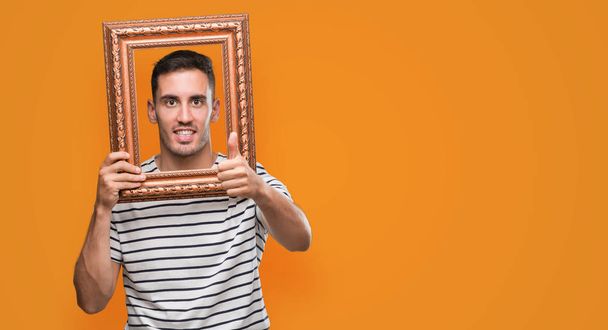 Handsome young man looking through vintage art frame happy with big smile doing ok sign, thumb up with fingers, excellent sign - 写真・画像