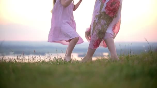 Cute little girl holding hands with mom stomps on the grass hill at sunset - Footage, Video