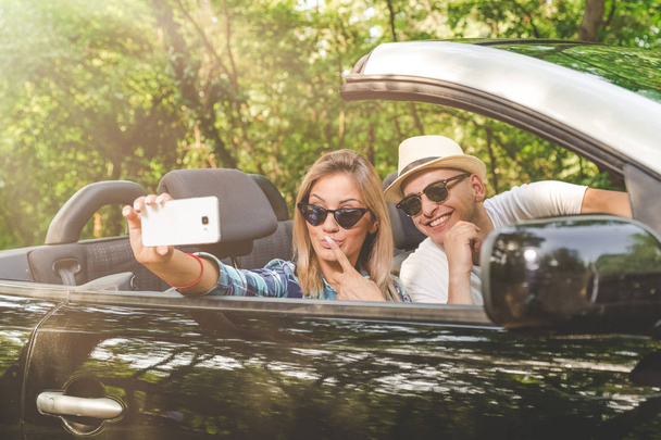 Young couple taking selfie with smartphone while sitting in convertible car. Travel and technology concepts. - Valokuva, kuva