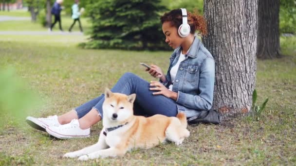 Attractive African American woman is listening to music with headphones and using smartphone sitting on grass in park while her dog is lying nearby eating grass. - Felvétel, videó
