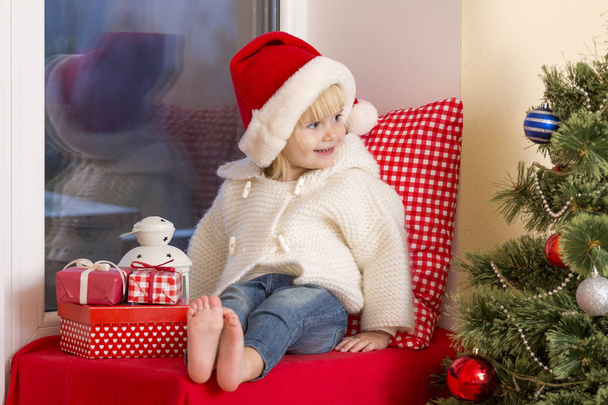 Happy family. A small girl in a santa's hat sits on a window sill in the window and waits for the saint. - Foto, Imagem
