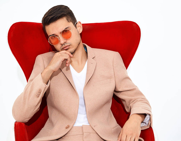 portrait of handsome fashion stylish hipster lumbersexual businessman model dressed in elegant light pink suit sitting on red chair on white background in studio. Metrosexual - Foto, immagini