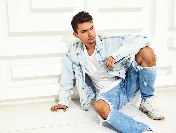 Portrait of handsome young model man dressed in jeans clothes sitting near white textured wall - Foto, Imagen