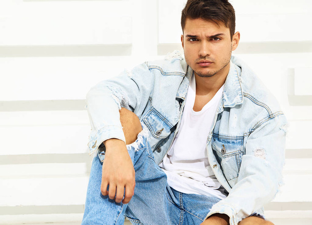 Portrait of handsome young model man dressed in jeans clothes sitting near white textured wall - Фото, изображение