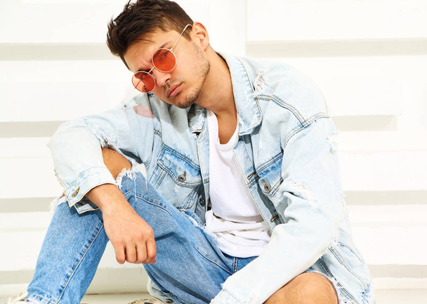 Portrait of handsome young model man dressed in jeans clothes sitting near white textured wall - Foto, afbeelding