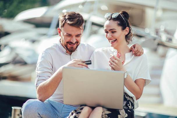 Beautiful romantic couple using laptop. Watching pictures on the laptop while traveling, by the harbor of a touristic sea resort with boats on background. - 写真・画像