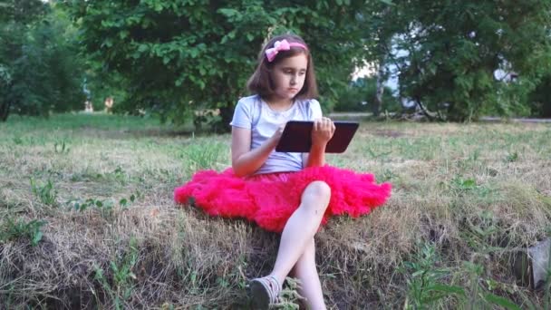little girl in a red skirt plays in a tablet on nature in the park sitting on the grass near the hollow - Filmati, video