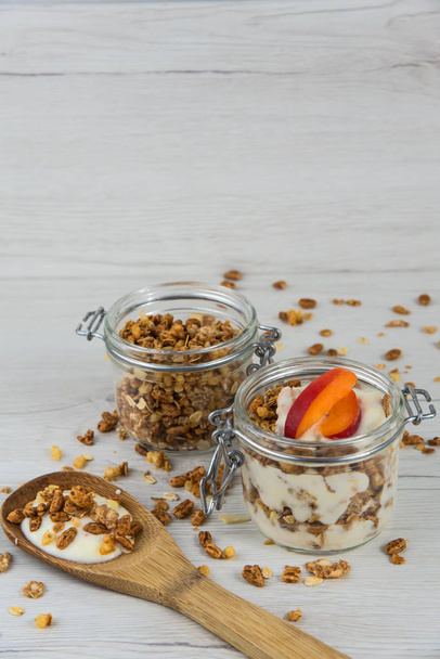 A jar tele granola és joghurt, fakanállal fikarcnyi granola és barack, fehér fa tábla - Fotó, kép