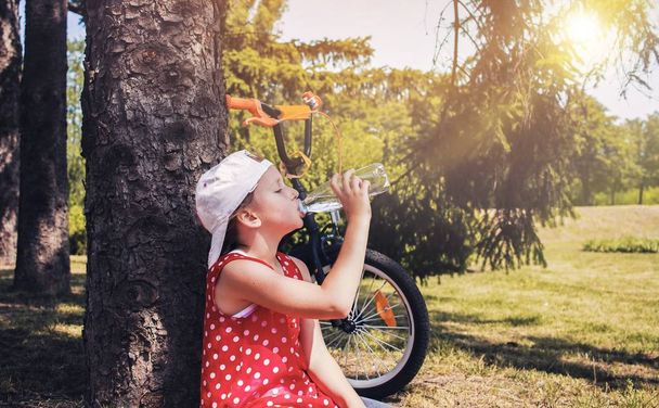 Roztomilá holka pije vodu z plastové láhve. Dítě pitnou vodu. horký letní den v parku. výlet na kole - Fotografie, Obrázek
