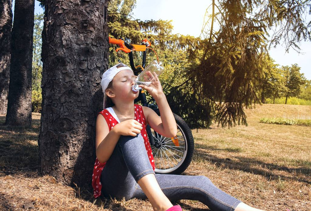 Ładna dziewczyna pije wodę z butelki z tworzyw sztucznych. Wody pitnej dziecko. gorący letni dzień w parku. Wycieczka rowerem - Zdjęcie, obraz