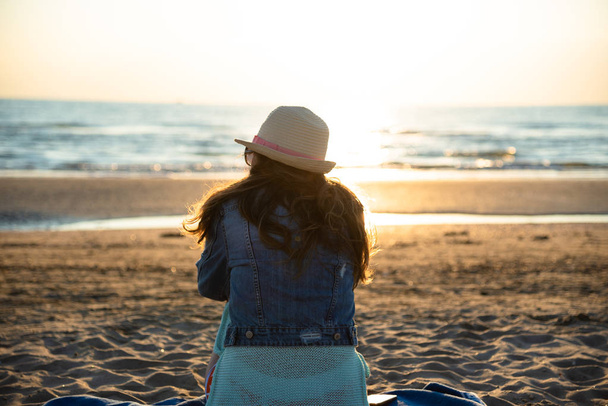 Tyttö istuu rannalla auringonnousun aikaan, ihaillen ja nauttien merituulesta
 - Valokuva, kuva