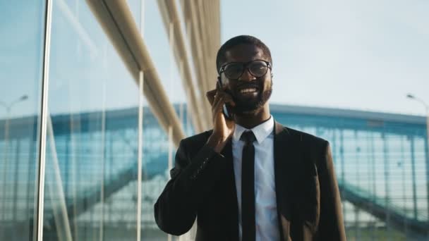Close up of attractive young African American businessman in a suit and tie walking slowly along a modern glass office building and talking on the mobile phone. Office worker heading to his workplace - Video, Çekim