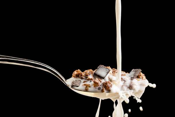 Milk splashing against silver spoon filled with cereals and chocolate over black - Фото, зображення