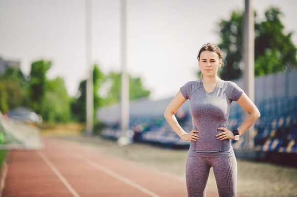 Portrait of a beautiful young caucasian woman with long hair in the tail and big breasts posing in gray sportswear standing training on a running stadium, a red rubber track in summer on a sunny day. - Valokuva, kuva