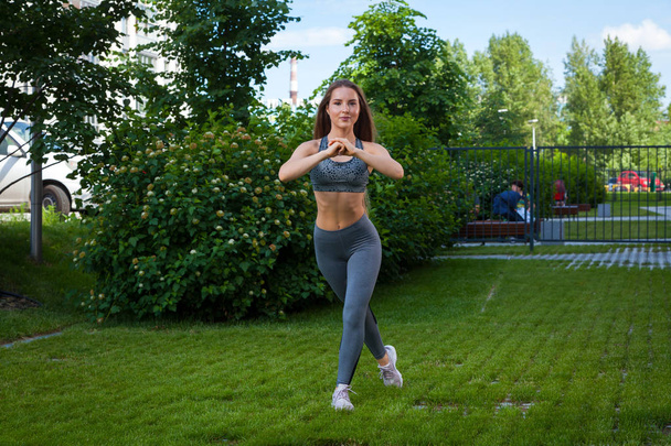 A dark-haired woman trainer in a sporty short top and gym leggings shows the correct technique of lunges for pumping the buttocks, hands in front of him on a summer day in a park on a green lawn - Photo, Image