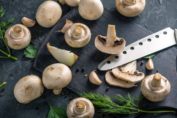 Fresh white champignons cut in half with a knife and the ingredients on the dark kitchen table. The concept of cooking mushroom dishes. Close up - Φωτογραφία, εικόνα