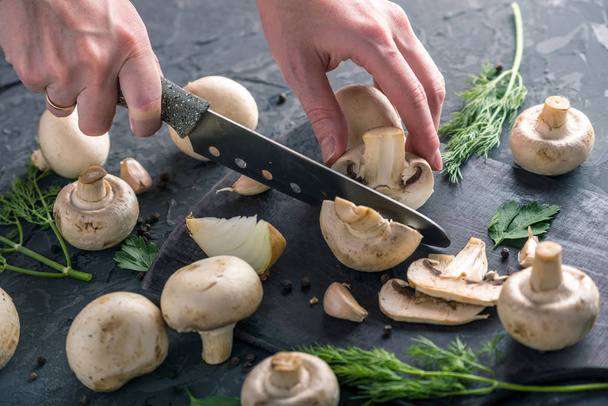 As mãos das mulheres estão cortando champinhons com uma faca na mesa da cozinha escura. O conceito de cozinhar pratos de cogumelos. Fechar
 - Foto, Imagem