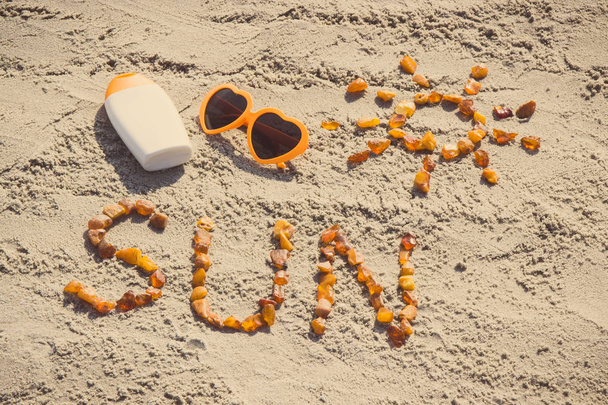 Inscription and shape of sun made of amber stones, sunglasses and sun lotion on sand at beach, concept of sunbathing, summer and vacation time - Photo, image