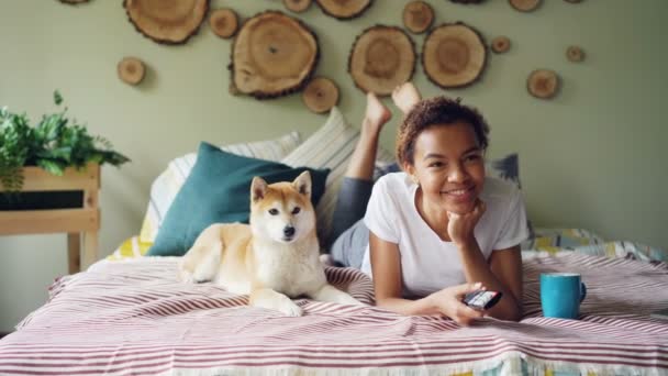 Cheerful mixed race student is watching TV holding remote and pressing buttons choosing television channels while her dog is moving on bed at home. - 映像、動画