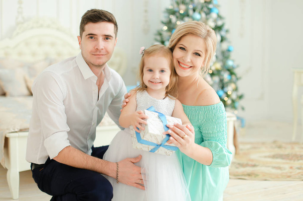 Mujer feliz y hombre sentado con hija pequeña manteniendo presente, árbol de Navidad en el fondo
. - Foto, imagen