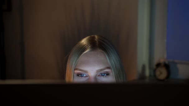 Una donna che sbircia fuori monitor del computer di notte. Vista ritagliare di bella femmina seduta di notte al computer e guardando la fotocamera sopra il monitor con finestra scura su sfondo sfocato - Filmati, video
