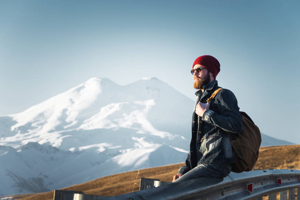 Bearded tourist hipster man in sunglasses with a backpack sitting on a roadside bump and watching the sunset against the background of a snow-capped mountain - Photo, image