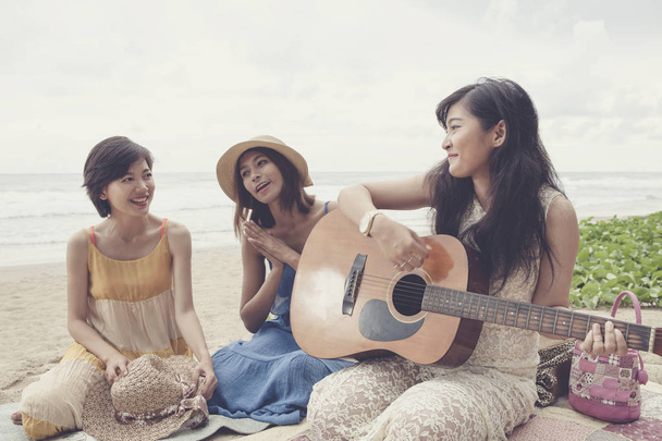 νεότερος γυναίκα της Ασίας φίλη ξεκούραστες διακοπές παίζει κιθάρα και να τραγουδήσει ένα τραγούδι στην παραλία της θάλασσας - Φωτογραφία, εικόνα