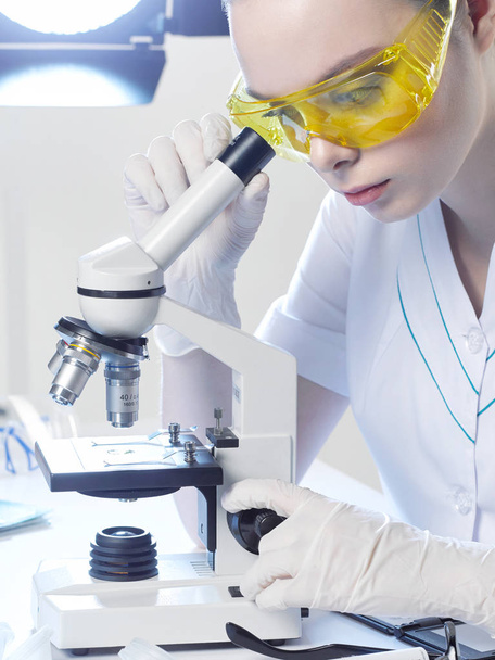 Jovem assistente de laboratório ou médico assistente, em óculos amarelos olha para um microscópio em um laboratório médico
. - Foto, Imagem