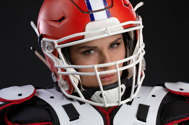 Porträt eines sexy attraktiven jungen Mädchens mit hellem Make-up in einem Sportoutfit für Rugby, mit Helm auf dem Kopf und starkem Blick nach vorne stehend auf schwarzem Hintergrund. Gleichberechtigung der Geschlechter - Foto, Bild