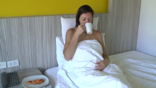 A woman lies in bed at a hotel, drinks coffee and eats pizza - Materiał filmowy, wideo