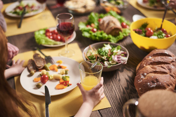 Close up of healthy dishes lying on table. Small girl is holding glass of orange juice while eating salad with vegetables - Photo, image
