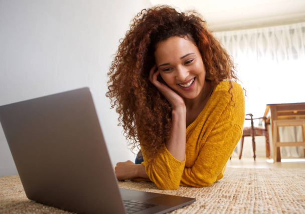Portrait of happy african american woman lying on floor looking at laptop computer  - 写真・画像