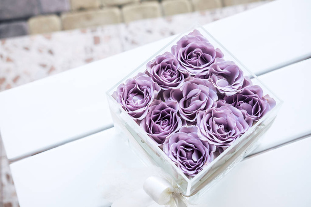 Pastel purple hued roses in clear acrylic crystal flower box. Square glass gift box. On white wooden table from above - Fotoğraf, Görsel