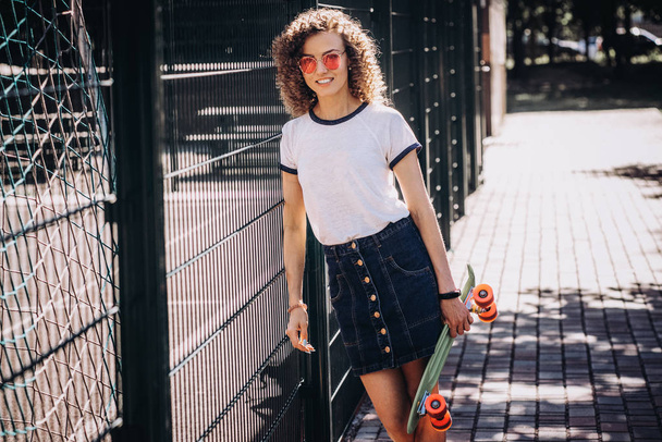 Skater curly woman in pink glasses outdoors with a skateboard - Fotoğraf, Görsel