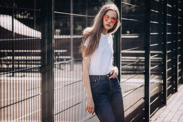 Beautiful young woman wearing jeans , white t-shirt, in pikn glasses standing on the street. Fashion photo near iron fence - Foto, imagen