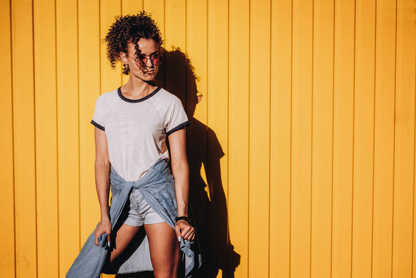 Portrait of a beautiful curly young woman in casual, wearing pink glasses, jeans and white t-shirt. smiling and while posing on yellow background - Fotó, kép