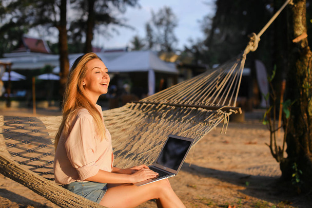 Freelancer feminino trabalhando com laptop e sentado na areia na rede de vime
. - Foto, Imagem