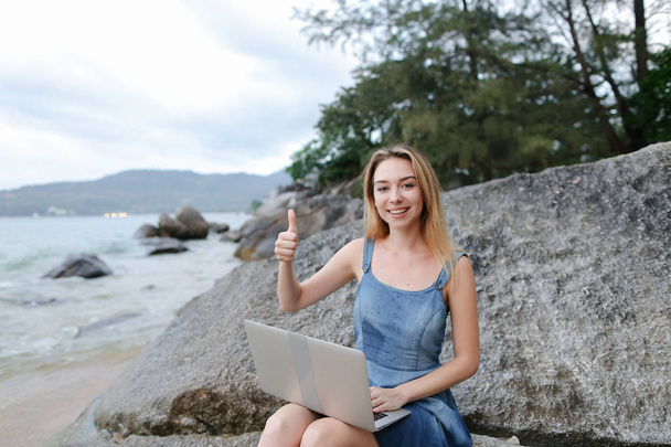 Молодая женщина с ноутбуком и сидя на камне у моря, пустой утренний пляж, показывая большие пальцы вверх
. - Фото, изображение