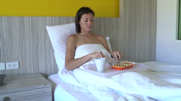 A woman is having breakfast with waffles and having coffee lying in bed in a hotel room - Imágenes, Vídeo