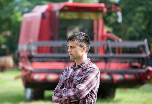 agricultor guapo de confianza de pie con los brazos cruzados delante de cosechadora en la granja
 - Foto, Imagen