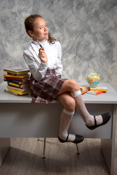 Das zappelige Schulmädchen in Schuluniform sitzt auf einem Tisch und lächelt glücklich auf einem hellgrauen Hintergrund. zurück zur Schule. Das neue Schuljahr. Kinderbildungskonzept. - Foto, Bild