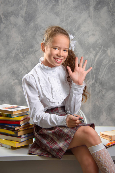 The fidget schoolgirl in school uniform sits on table and smiles happily on a light gray background. Back to school. The new school year. Child education concept. - Zdjęcie, obraz