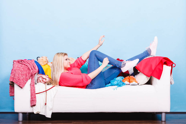 Clothing dilemmas concept. Happy lazy woman lying on messy couch with piles of clothes - 写真・画像