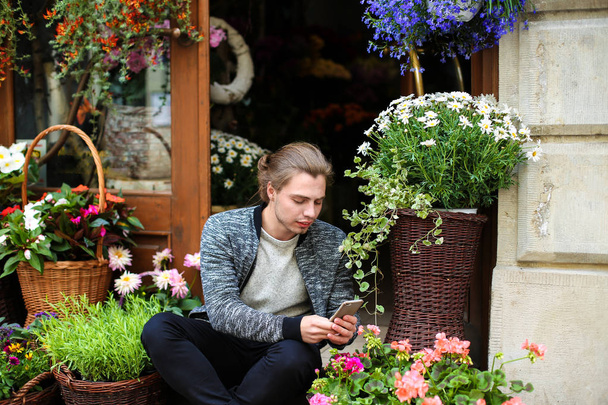 Junger Kaukasier plaudert per Smartphone und sitzt im Blumenladen in der Nähe von Blumensträußen in Körben. - Foto, Bild
