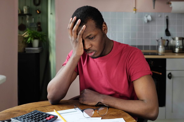 Nuoret turhautunut onneton väsynyt afrikkalainen mies taloudellisia ongelmia, istuu keittiön pöydässä paperit, laskin ja kasvojen kämmen ele
 - Valokuva, kuva